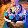 baby, smiling, walker, toy, indoor, child, happy, person, living_room, carpet, furniture, seat, bib, head, face, playtime, cushion, spiderman, colorful, floor