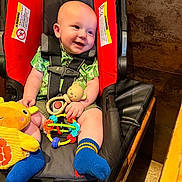 Jaxon joined the competition — help win amazing prizes! baby, infant, car_seat, child, toy, blue_socks, green_shirt, smile, happy, indoor, seatbelt, plush_toy, colorful_toy, red_padding, face, person, cute, young_child, sitting, comfort