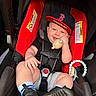 Jaxon is registered to the contest to win money with this photo: baby, car_seat, child, toy, cap, black_shirt, gray_shorts, blue_striped_socks, black_shoes, smiling, face, hand, footwear, seat_belt, infant, indoor, cute, childhood, comfort, red_car_seat