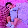baby, child, smiling, curly_hair, onesie, socks, pink, purple, plush, blanket, couch, happy, infant, person, cute, soft_texture, lying_down, cozy, indoors, portrait