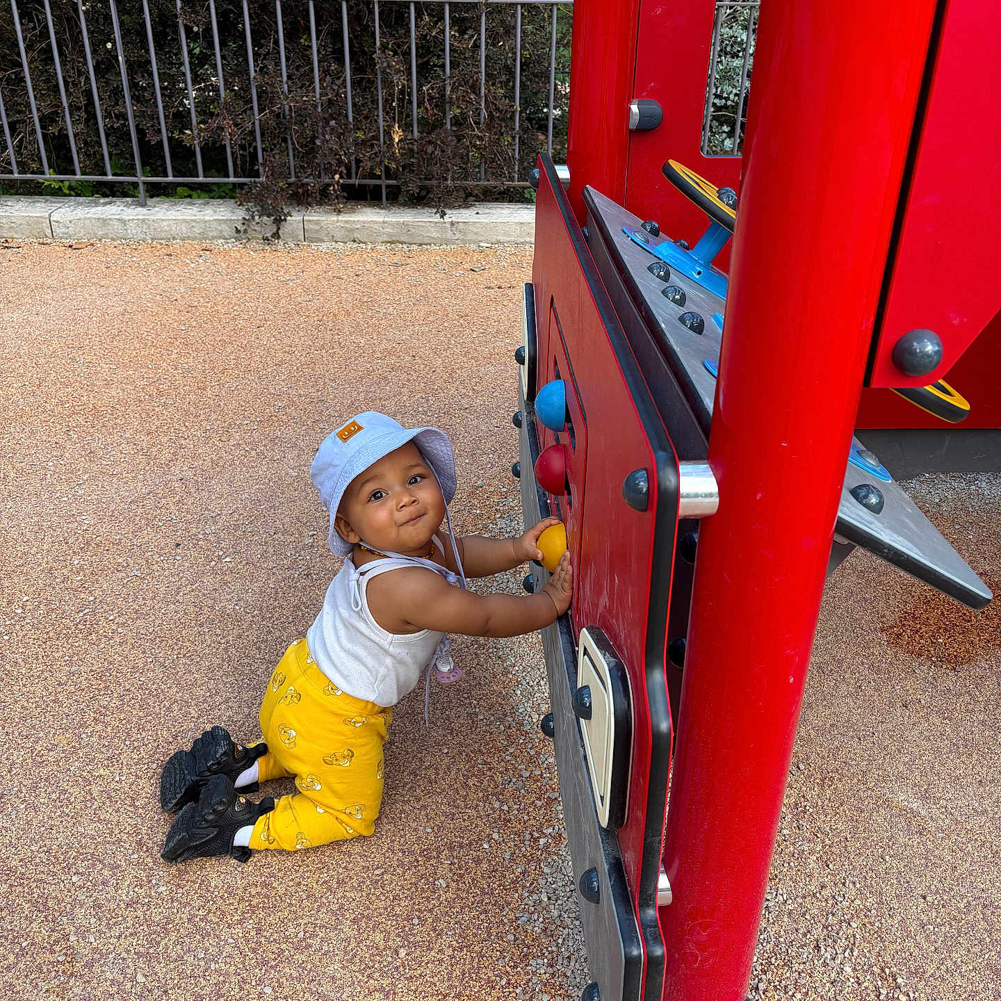 Thaysse participe au concours pour gagner de l'argent avec cette photo : black_shoes, child, curious, daylight, fence, footwear, greenery, happy, hat, kneeling, outdoor, person, play, play_structure, playground, red, sunny, toddler, toy, yellow_pants