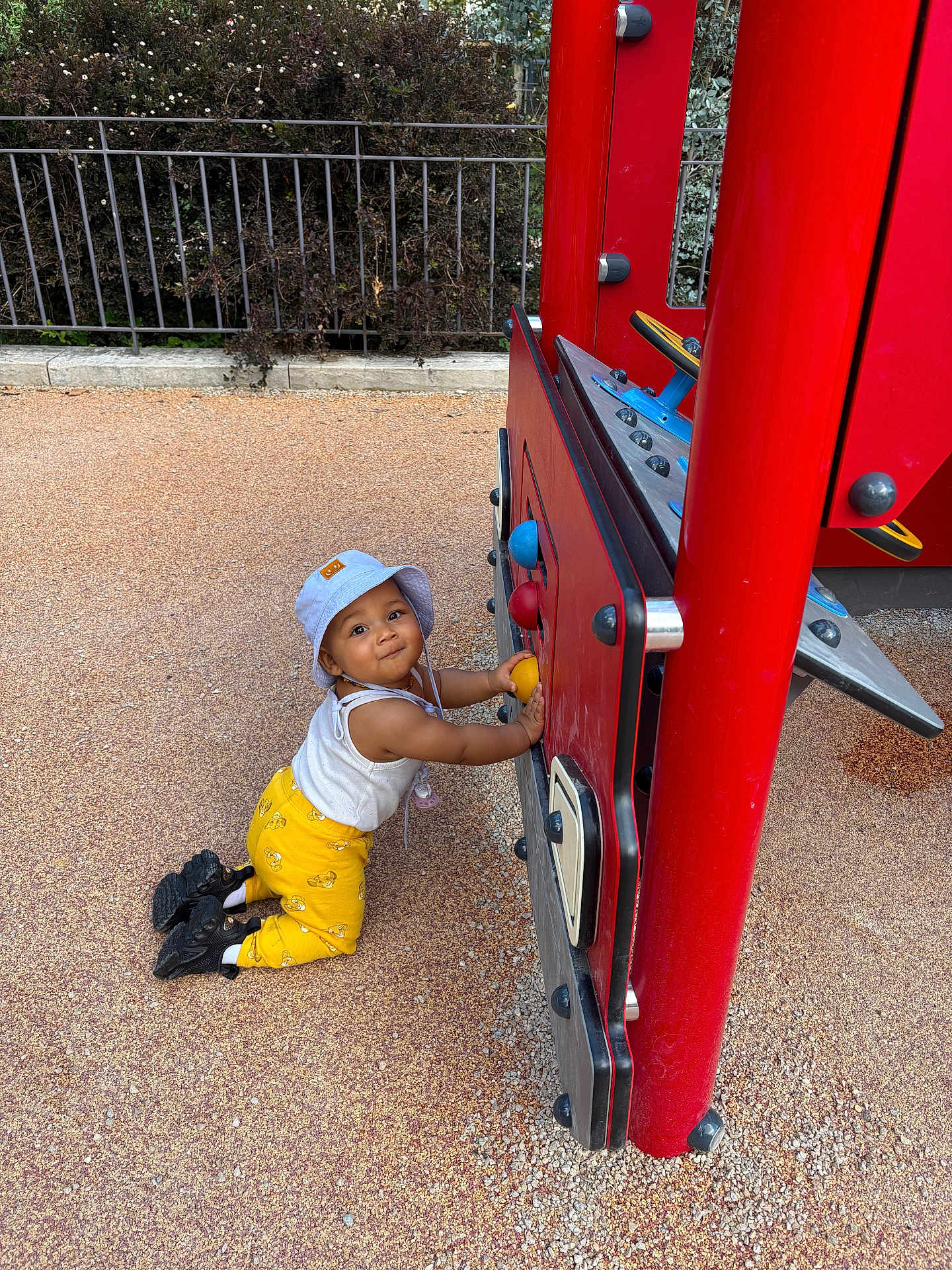 Thaysse participe au concours pour gagner de l'argent avec cette photo : toddler, child, playground, red, hat, yellow_pants, kneeling, outdoor, play_structure, toy, curious, fence, greenery, footwear, black_shoes, sunny, daylight, play, person, happy