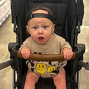 Greyson is registered to the contest to win money with this photo: baby, infant, stroller, stroller_handle, cap, backwards_cap, blue_eyes, surprised_expression, gripping, smiley_shirt, onesie, hands, legs, seat, portrait, cute, indoor, shopping_aisle, bokeh, child
