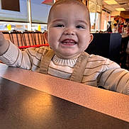 Ethan joined the competition — help win amazing prizes! baby, smiling, child, indoor, restaurant, table, overalls, sweater, happy, face, person, dining, light, window, people, seat, furniture, casual, childhood, portrait