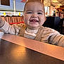 baby, smiling, child, indoor, restaurant, table, overalls, sweater, happy, face, person, dining, light, window, people, seat, furniture, casual, childhood, portrait