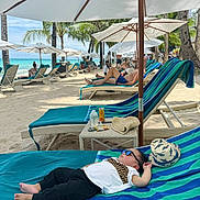 Thirdy is registered to the contest to win money with this photo: child, toddler, beach, lounger, umbrella, sunglasses, hat, sand, ocean, palm_tree, people, relaxing, vacation, sunshade, summer, blue_sky, towel, drink, table, holiday