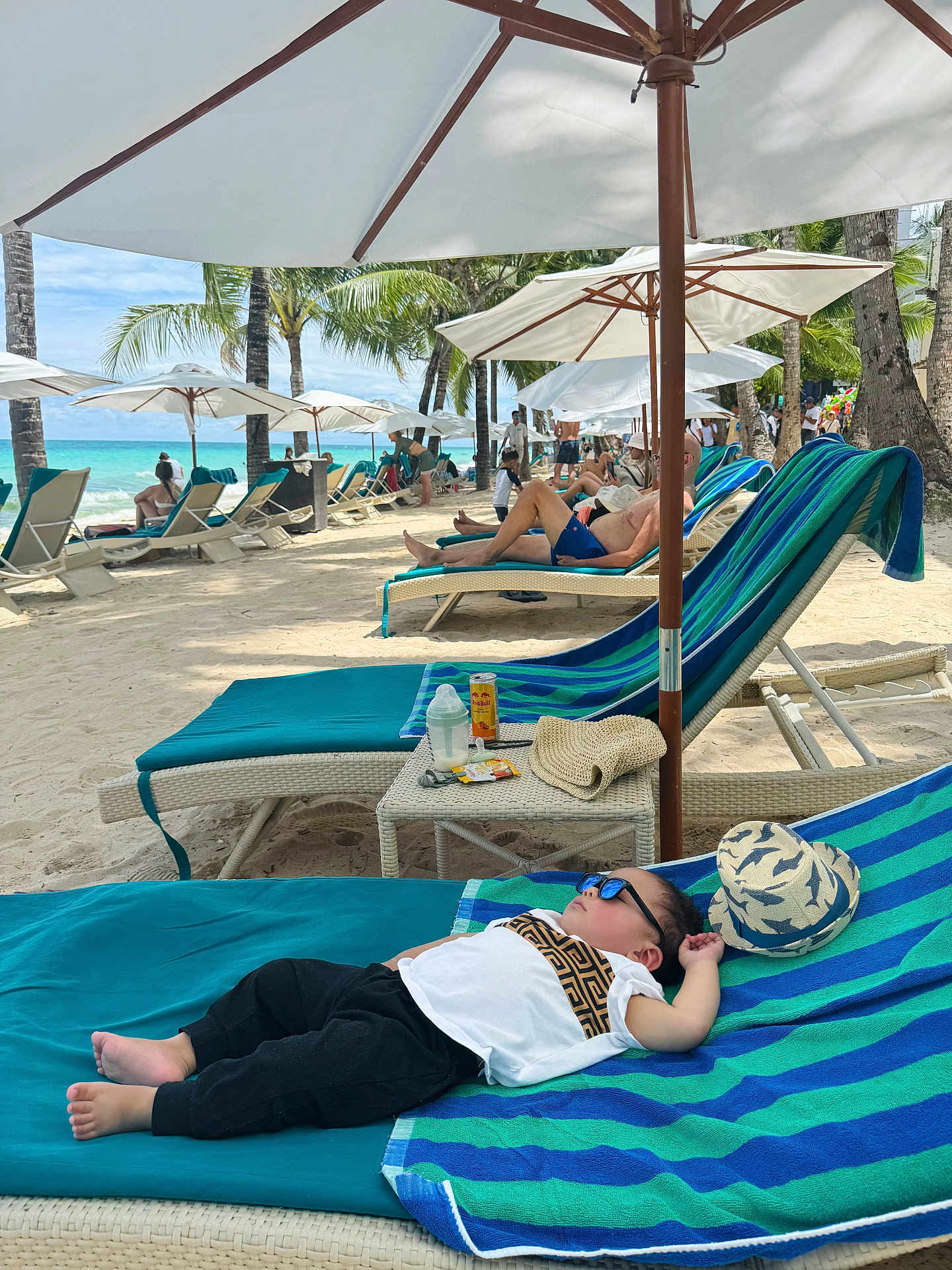 Thirdy is registered to the contest to win money with this photo: child, toddler, beach, lounger, umbrella, sunglasses, hat, sand, ocean, palm_tree, people, relaxing, vacation, sunshade, summer, blue_sky, towel, drink, table, holiday