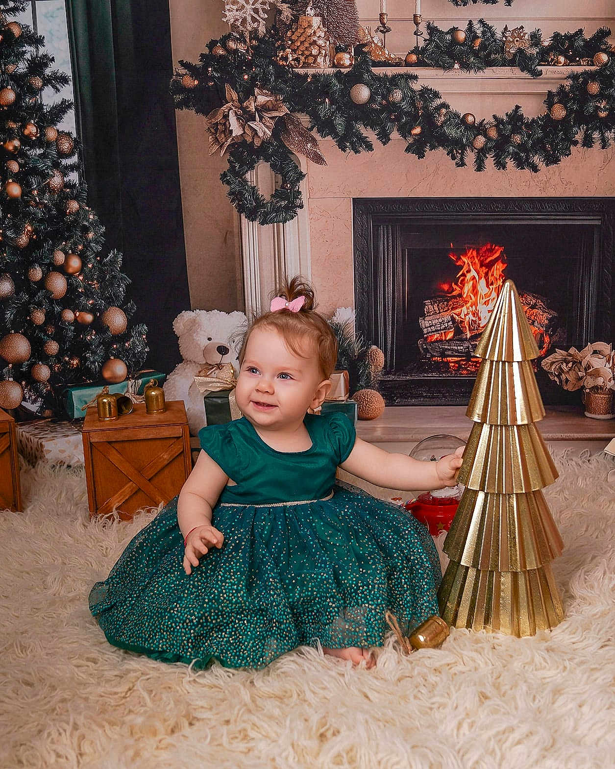 Iris is registered to the contest to win money with this photo: baby, child, dress, green_dress, smiling, sitting, christmas_tree, christmas_decorations, fireplace, teddy_bear, gift_boxes, golden_tree, holiday, festive, indoors, carpet, happy, cute, portrait, celebration