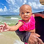 baby, child, person, beach, ocean, sky, clouds, water, sand, hand, arm, pink_clothing, outdoor, sunny, portrait, expression, holding, adult, summer, vacation