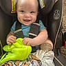 baby, smiling, car_seat, blanket, bottle, infant, child, happy, blue_shirt, safety_harness, indoor, cute, clothing, face, person, seat, baby_toy, car_seat_warning_label, holding, comfort