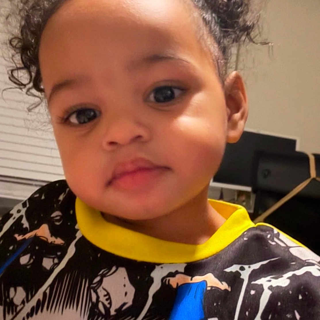 Leilani joined the competition — help win amazing prizes! black_hair, blouse, casual_clothing, child, closeup, curly_hair, cute, expression, eyebrows, eyelashes, face, home, indoor, person, portrait, skin, smiling, superhero_shirt, toddler, young_child