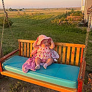 Esabella is registered to the contest to win money with this photo: baby, child, swing, bench, hat, pink_clothing, outdoor, grass, field, sunset, wood, cushion, rural, nature, person, socks, plush_blanket, daylight, fence, house