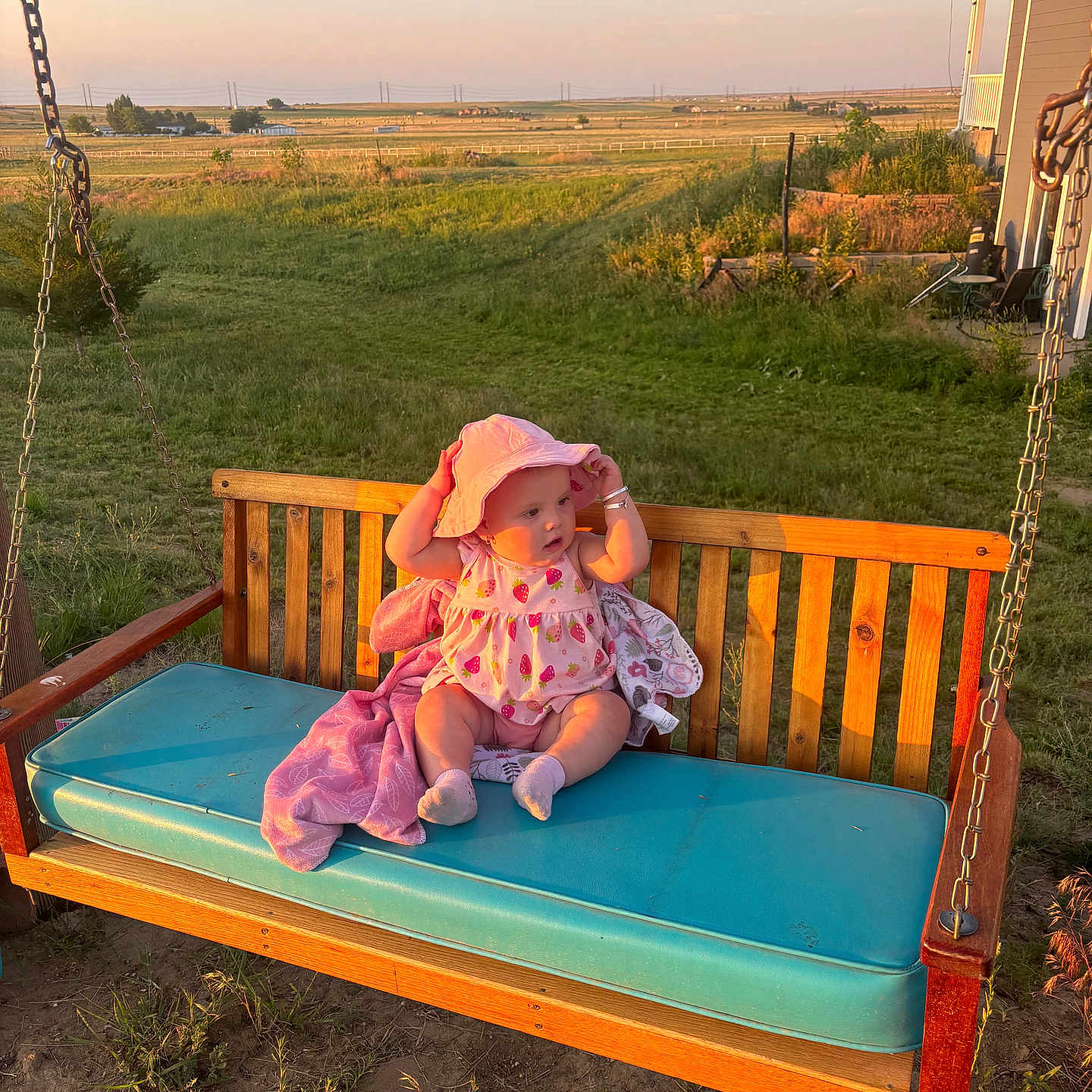 Esabella is registered to the contest to win money with this photo: baby, bench, child, cushion, daylight, fence, field, grass, hat, house, nature, outdoor, person, pink_clothing, plush_blanket, rural, socks, sunset, swing, wood