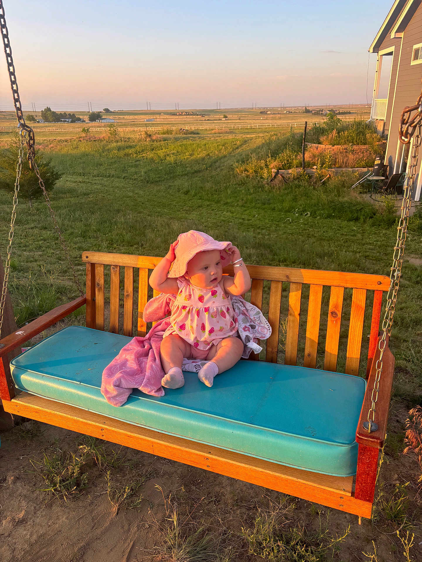 Esabella is registered to the contest to win money with this photo: baby, child, swing, bench, hat, pink_clothing, outdoor, grass, field, sunset, wood, cushion, rural, nature, person, socks, plush_blanket, daylight, fence, house