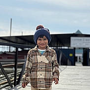 Agastya is registered to the contest to win money with this photo: child, boy, plaid_shirt, beanie, hat, smiling, outdoor, pier, wooden_deck, water, cloudy_sky, casual_clothing, standing, young, person, daylight, cool_weather, happy, portrait, background_blur