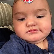 Emily joined the competition — help win amazing prizes! baby, child, infant, portrait, close_up, face, serious_expression, indoors, clothing, blue_shirt, sticker, forehead, eyes, nose, cheeks, couch, blanket, seated, selfie, cute