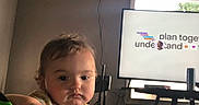 Emily is registered to the contest to win money with this photo: baby, blanket, brown_hair, child, couch, face, indoors, living_room, person, photograph, pointing, portrait, presentation, screen, serious, shirt, sitting, television, toddler, window