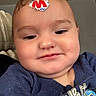 baby, child, infant, portrait, close_up, face, eyes, cheeks, forehead_sticker, sticker, blue_shirt, graphic_shirt, sitting, indoors, pillow, couch, soft_lighting, looking_at_camera, serious_expression, photograph