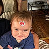 Emily is registered to the contest to win money with this photo: child, toddler, baby, portrait, face, smiling, forehead_sticker, blue_shirt, red_wings, adult, arm, indoor, living_room, wood_floor, coffee_table, couch, blanket, casual, close_up, family
