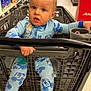 toddler, child, shopping_cart, pajamas, mickey_mouse, blue_clothing, supermarket, aisle, face, expression, hand, floor, product_shelves, plastic_cart, indoor, baby, person, retail, store, curious
