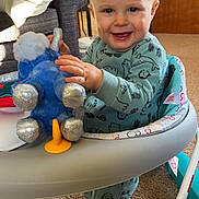 Benjamin is registered to the contest to win money with this photo: baby, child, smiling, pajamas, plush_toy, walker, indoor, carpet, furniture, living_room, toy, cute, standing, happy, person, footwear, animal_prints, cozy, home, infant