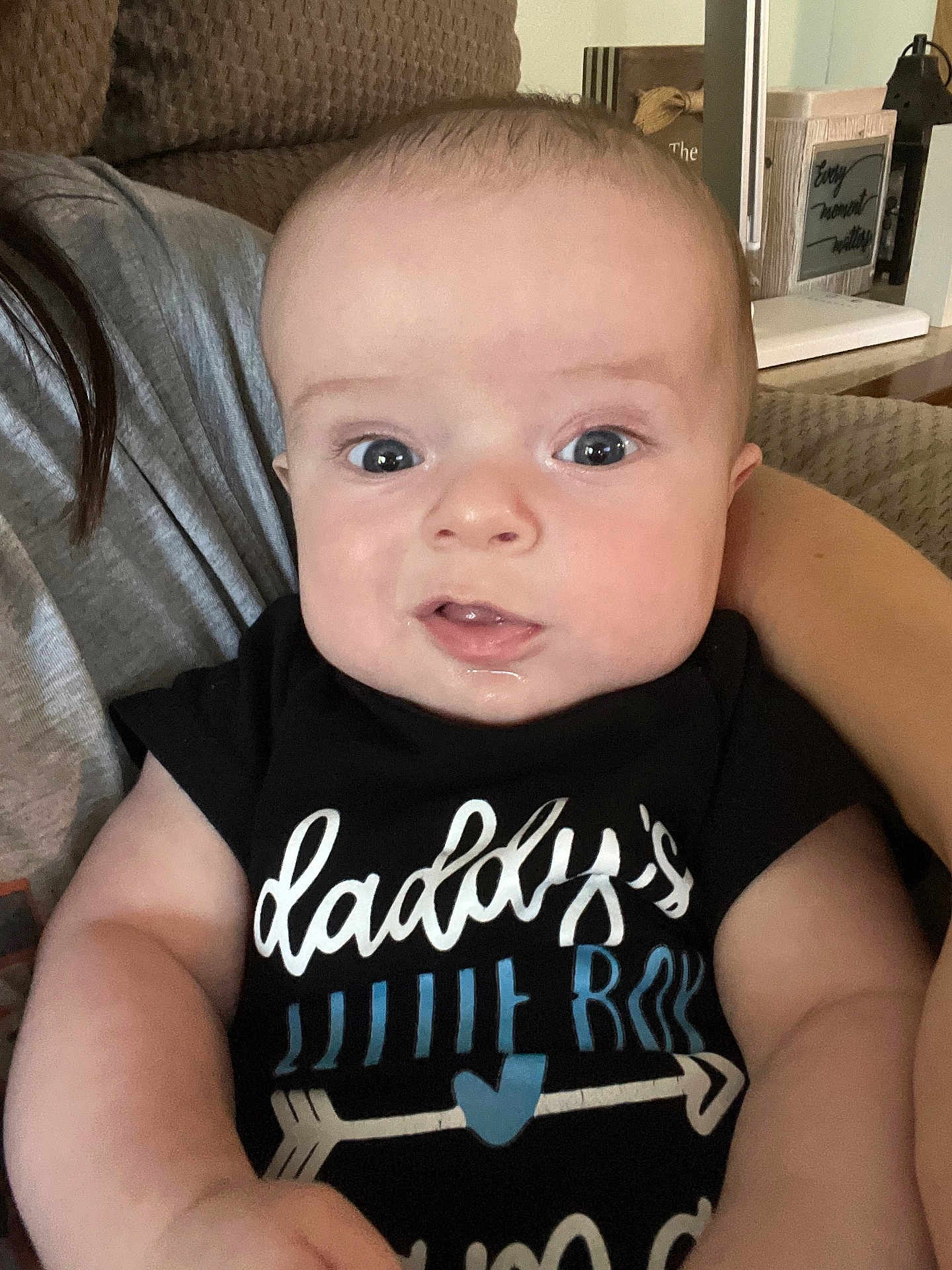 James is registered to the contest to win money with this photo: baby, infant, person, face, arm, clothing, black_shirt, text_on_clothing, drool, indoor, couch, furniture, home_decor, curious, skin, head, young_child, holding, close_up, seated