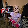 toddler, child, smiling, purple_dress, bottle, indoor, living_room, carpet, furniture, photograph, toy, sneakers, person, happy, face, hair, table, box, blanket, home