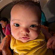 Rayana joined the competition — help win amazing prizes! baby, child, face, eyes, hand, yellow_clothing, indoor, portrait, curly_hair, person, closeup, infant, cute, holding, skin, expression, looking, people, childhood, warm_lighting