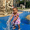 child, girl, toddler, outdoor, waterpark, fountain, water_splash, striped_dress, colorful_clothing, palm_tree, rock, waterfall, sunny, summer, barefoot, smiling, playing, portrait, pool, recreation