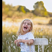 Sopho joined the competition — help win amazing prizes! child, girl, laughing, white_chair, grass, field, outdoor, nature, sunlight, blonde_hair, casual_clothing, blue_jeans, white_shirt, happy, portrait, sitting, daylight, smile, greenery, trees