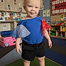 toddler, child, indoor, playroom, blue_shirt, black_shorts, barefoot, curly_hair, smiling, educational_posters, toys, play_block, classroom, colorful, pumpkins, furniture, person, cute, happy, young_child