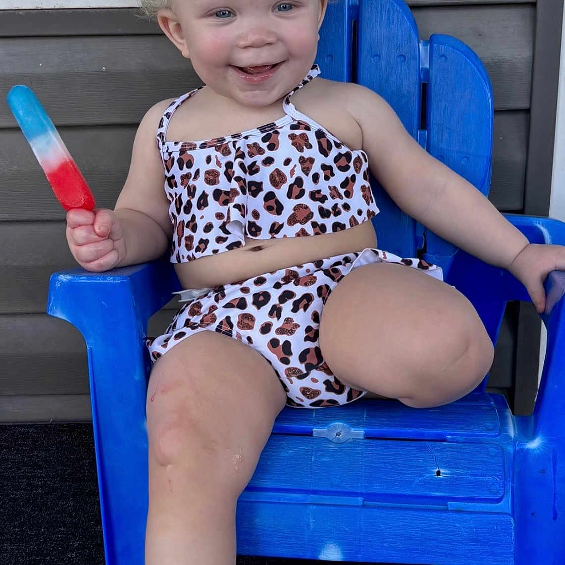 Camilla is registered to the contest to win money with this photo: toddler, child, smiling, curly_hair, leopard_print, two_piece_outfit, blue_chair, popsicle, red_white_blue, porch, home, sitting, happy, summer, outdoor, plastic_chair, cute, fun, playful, young_child