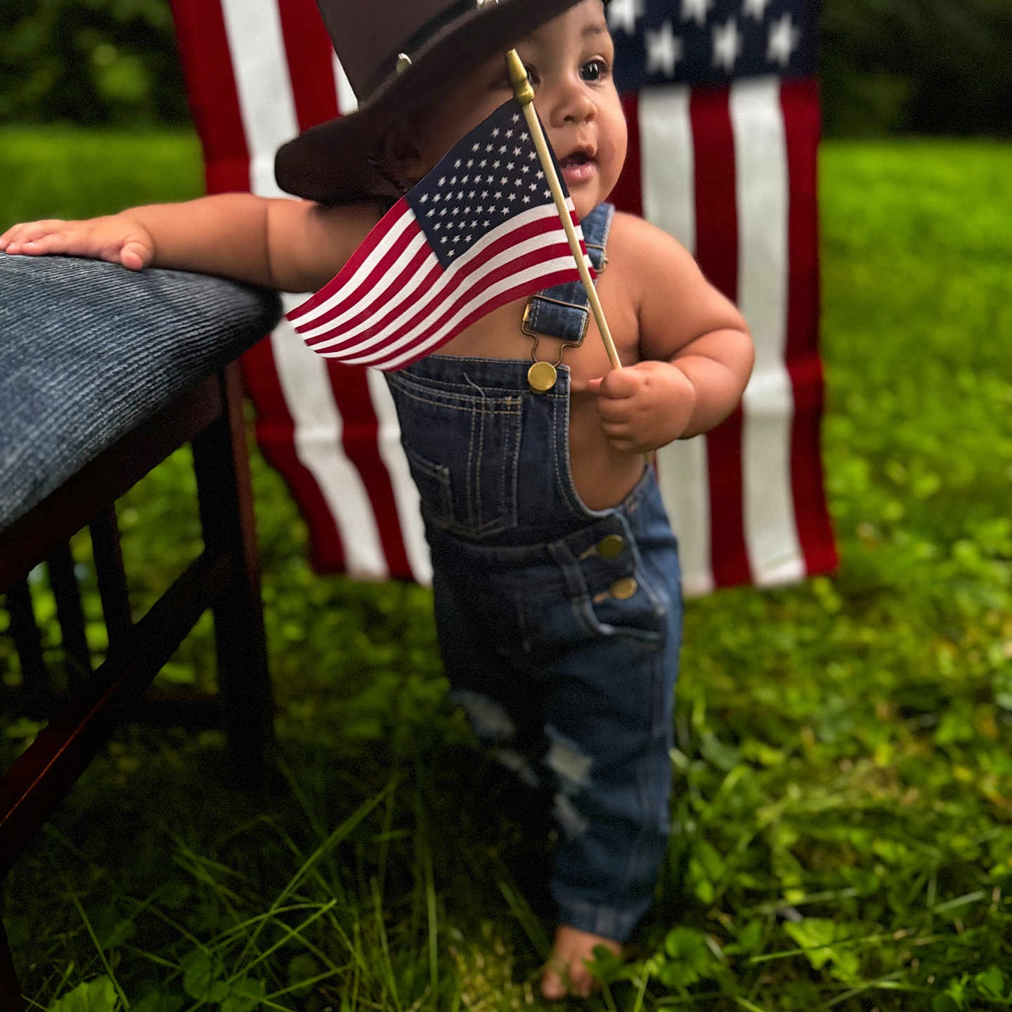 Amari joined the competition — help win amazing prizes! toddler, child, american_flag, cowboy_hat, denim_overalls, grass, outdoor, barefoot, chair, patriotic, flag, person, baby, portrait, summer, greenery, cute, standing, holiday, nature