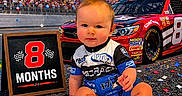 Riley is registered to the contest to win money with this photo: automobile, baby, checkered_flag, child, confetti, finish_line, grandstand, infant, number_eight, outdoor, photography, portrait, race_car, race_track, racing, racing_suit, red_car, sitting, smiling, stadium