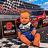 Riley is registered to the contest to win money with this photo: baby, infant, child, sitting, smiling, portrait, racing, race_car, race_track, checkered_flag, finish_line, confetti, stadium, grandstand, automobile, red_car, racing_suit, number_eight, outdoor, photography