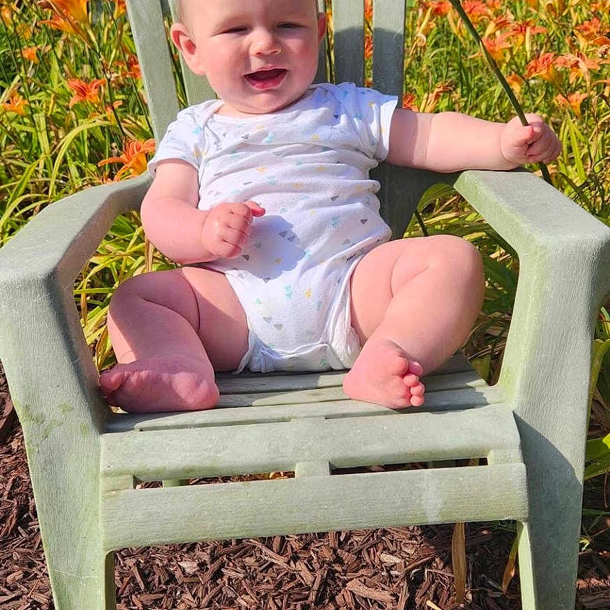 Riley joined the competition — help win amazing prizes! baby, child, outdoor, flower, lily, green_chair, smiling, sunlight, nature, plants, grass, summer, happy, person, one_piece_outfit, barefoot, sitting, garden, daylight, flora