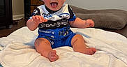 Riley joined the competition — help win amazing prizes! baby, blue_clothing, child, couch, curtains, furniture, happy, home, indoor, infant, living_room, open_mouth, portrait, racing_outfit, sitting, smiling, waving, white_blanket, window, wood_floor
