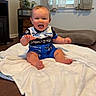 Riley joined the competition — help win amazing prizes! baby, child, infant, sitting, smiling, happy, open_mouth, waving, portrait, racing_outfit, blue_clothing, white_blanket, indoor, living_room, window, curtains, couch, furniture, wood_floor, home