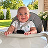 baby, child, high_chair, smiling, happy, portrait, outdoor, backyard, garden, lawn, trees, blue_sky, sunlight, white_fence, flowers, brick_wall, infant, sitting, harness, table