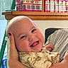 baby, child, infant, smiling, happy, tongue_out, portrait, close_up, indoor, home, being_held, caregiver, arm, face, eyes, bald_head, short_sleeves, clothing, shelf, storage_boxes