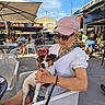 cafe, chairs, dog, elderly_person, holding_dog, jack_russell_terrier, outdoor, palm_tree, patio, pet, pink_cap, restaurant, seated, small_dog, street, sunglasses, tables, umbrella, white_shirt, woman