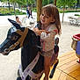 amusement_ride, carousel, child, daylight, fun, girl, happy, horse, jacket, outdoor, park, people, playground, recreation, shorts, smiling, sneakers, summer_clothing, trees, wooden_floor