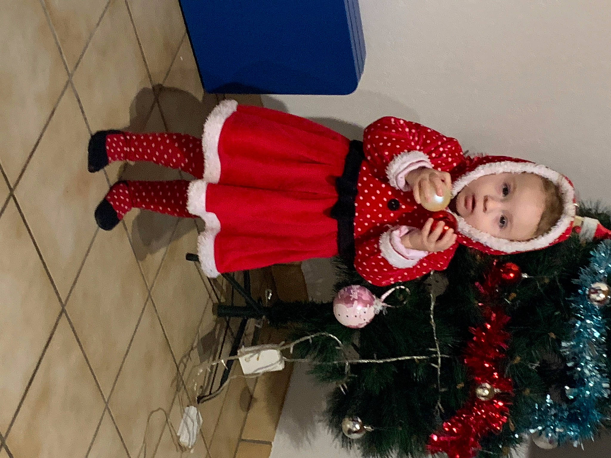 Lyliana a rejoint le concours — aidez-le/la à gagner de superbes lots ! carmine, christmas_decoration, christmas_eve, christmas_tree, costume, event, flooring, fun, holiday, human_leg, lap, pattern, person, plant, red, room, santa_claus, smile, toddler, tradition