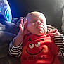 baby, infant, sleeping, tongue_out, red_clothing, striped_sleeves, smiley_face, text_on_clothing, blanket, soft_texture, hand_gesture, game_controller, glowing_lights, indoor, cute, child, portrait, resting, playful, young