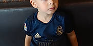 Lisandro participe au concours pour gagner de l'argent avec cette photo : child, boy, sitting, table, restaurant, booth, casual_clothing, jeans, short_hair, indoor, portrait, looking_away, navy_jersey, sportswear, adidas_logo, real_madrid_jersey, hands_on_bench, wood_paneling, young_person, calm_expression