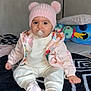 baby, child, pacifier, pink_hat, pom_poms, jacket, floral_pattern, socks, blanket, plush_toys, indoor, cute, seated, cozy_clothing, patterned_blanket, soft_toys, infant, person, warm_clothes, portrait