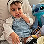 baby, child, girl, headband, bow, denim_overalls, couch, toy, plush_toy, stitch, hand, fingers, white_shirt, cute, indoor, smiling, person, infant, soft_toy, home