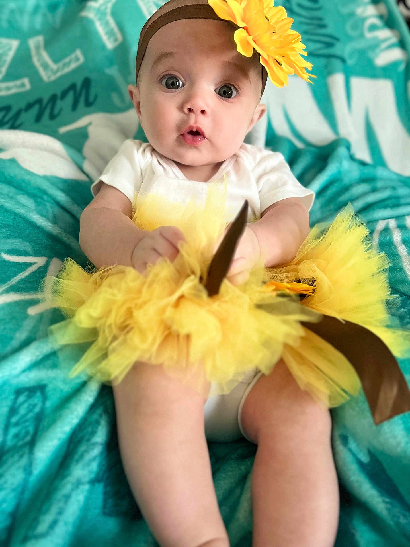 Jazlynn is registered to the contest to win money with this photo: baby, infant, tutu, headband, flower, yellow, white_clothing, blanket, teal, sitting, cute, portrait, child, wide_eyes, expression, soft_fabric, indoors, person, young_child, closeup