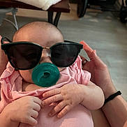 Jazlynn joined the competition — help win amazing prizes! baby, sunglasses, pacifier, pink_clothing, hands, infant, indoor, person, adult_hand, cute, cozy, playful, portrait, child, human, closeup, soft_lighting, casual, relaxed, home