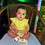 baby, child, yellow_dress, white_sandals, wicker_chair, cushion, holding_object, outdoor, night, garden_lights, greenery, plants, curious, portrait, cute, infant, sitting, round_sign, colorful_lights, festive